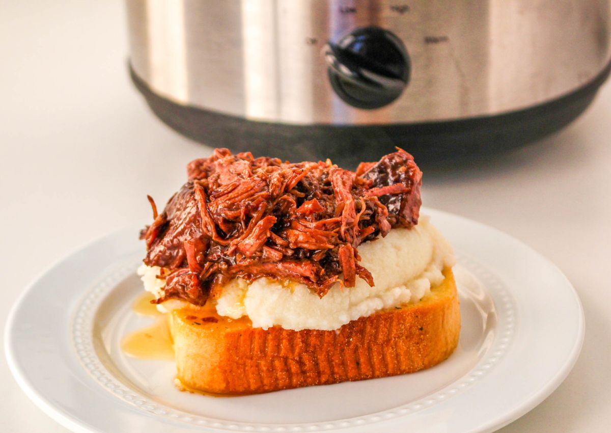A slice of toasted bread topped with creamy mashed potatoes and savory pulled beef, with a slow cooker in the background.