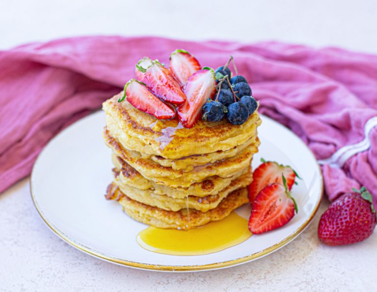 A stack of pancakes topped with sliced strawberries, blueberries, and syrup on a white plate. A pink cloth is in the background, and a whole strawberry is placed beside the plate.